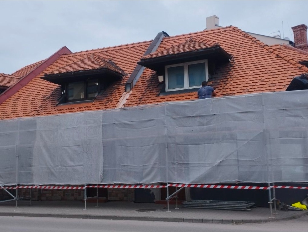 Ceglasty dach z oknami połaciowymi w trakcie prac. Widoczne rusztowania z siatką ochronną i pracownik przy oknie. Ujęcie z poziomu ulicy.