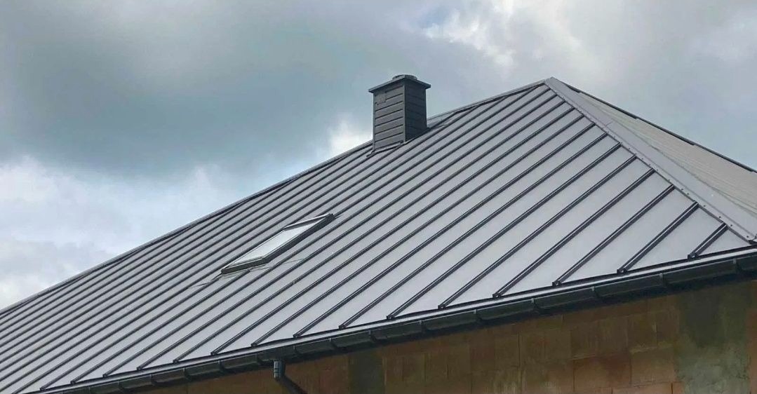 Szary dach z blachy na budynku z cegły, widoczny komin i okno dachowe. Pochmurne niebo w tle. Ujęcie z dołu, podkreślające geometrię dachu.