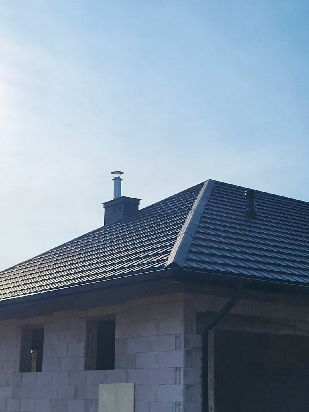 Nowy, grafitowy dach z dachówki ceramicznej na surowym budynku z pustaków, z widocznym kominem i rynnami w słoneczny dzień. Dachówka ułożona w karo.