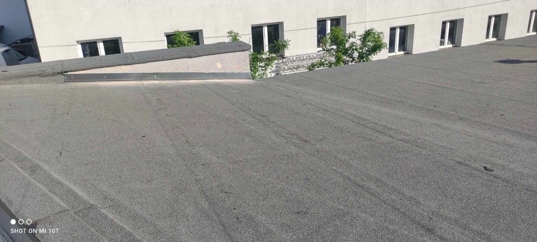 Szary, płaski dach pokryty papą na tle białego budynku z oknami. Widoczne detale łączeń papy i obróbek blacharskich. Ujęcie z góry, w słoneczny dzień.