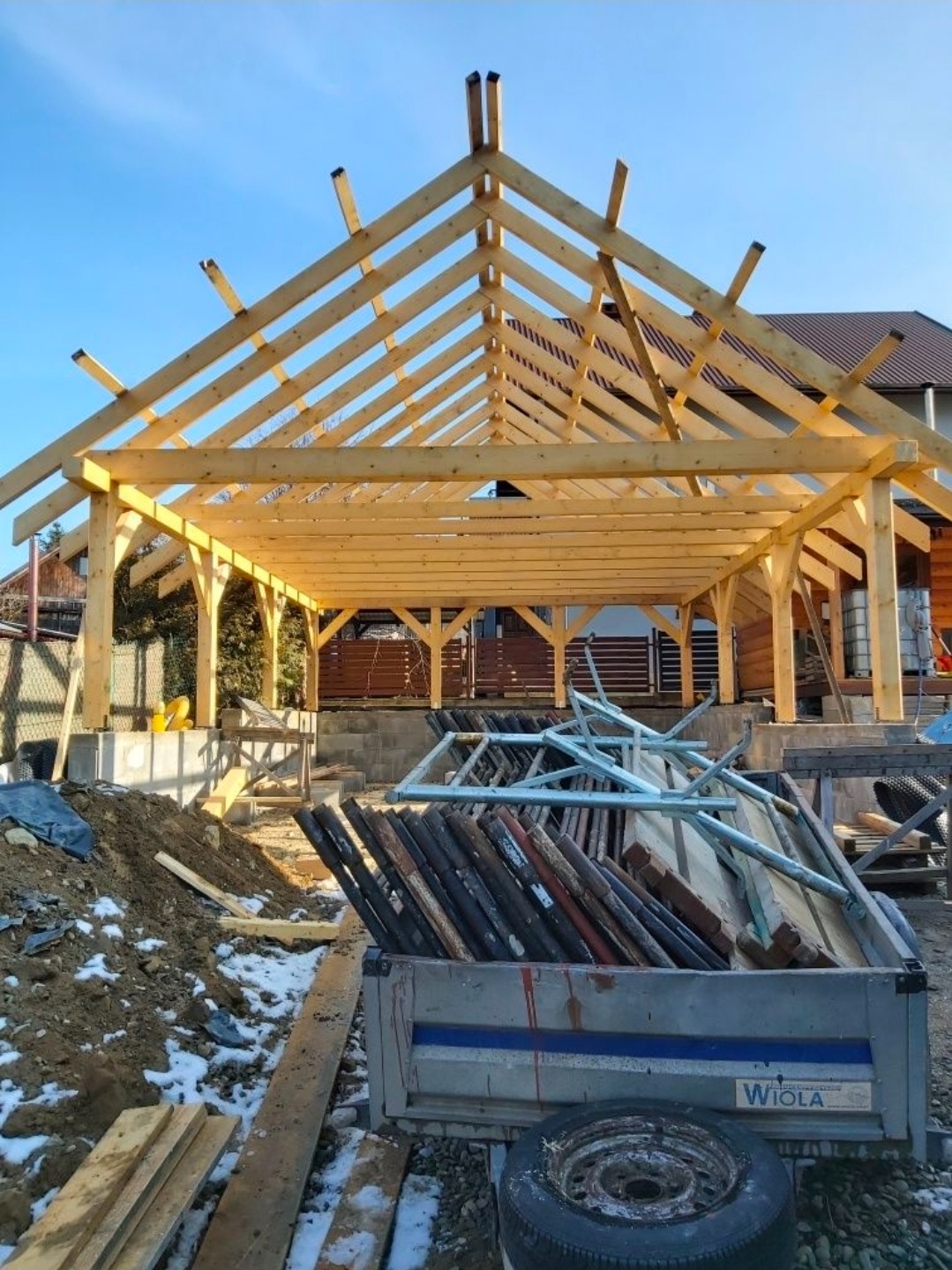 Drewniana konstrukcja dachu w trakcie budowy, widok z dołu na jasne niebo. W tle dom z brązowym dachem. Przyczepka z materiałami budowlanymi na pierwszym planie.