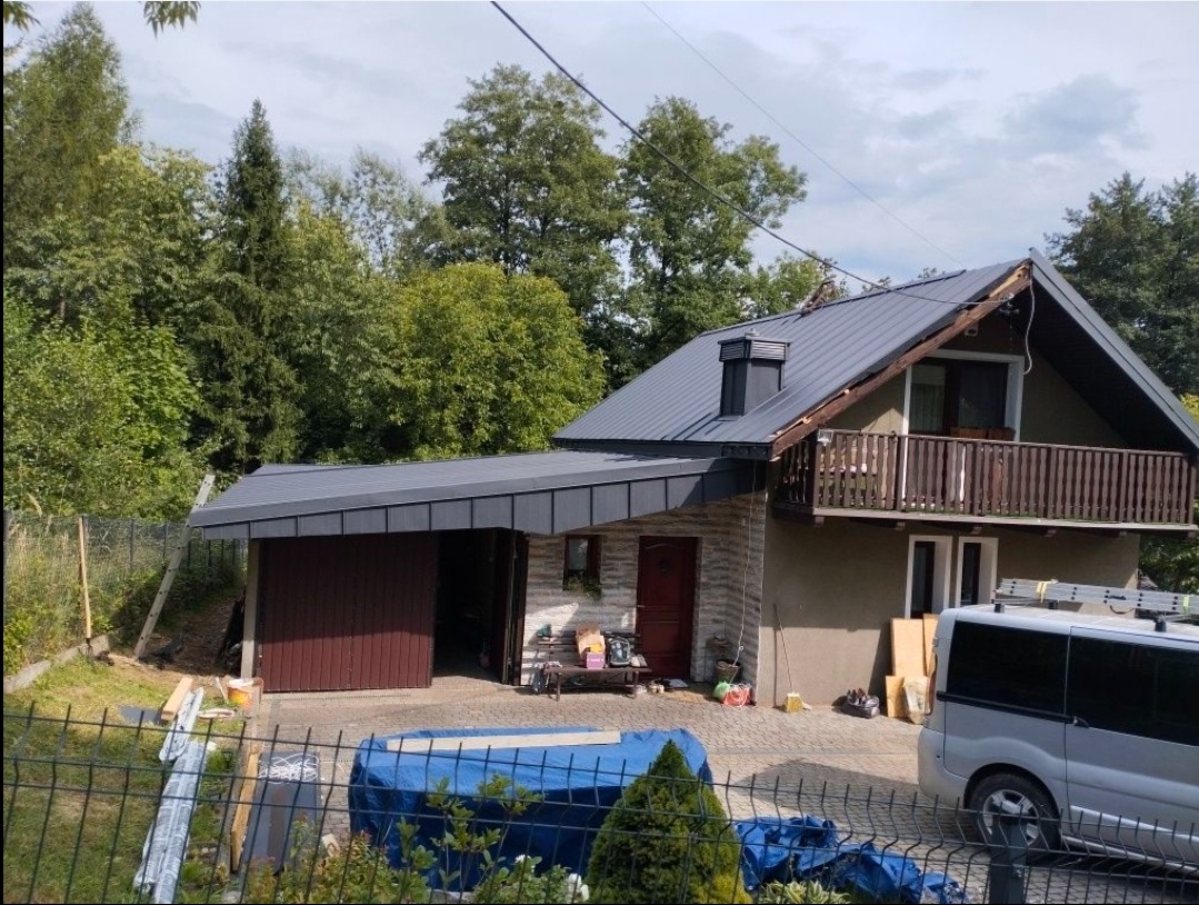 Dom z szarą blachą na dachu i dobudowanym garażem. Widoczne obróbki blacharskie i elementy wykończeniowe. Na podjeździe zaparkowany samochód dostawczy.