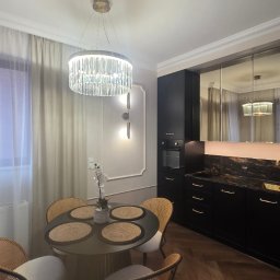 Elegancka, czarna kuchnia z marmurowym blatem i lustrzanymi szafkami wiszącymi. Okrągły stół z rattanowymi krzesłami pod kryształowym żyrandolem.