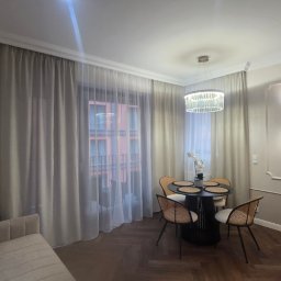 Eleganckie wnętrze: salon z sofą, okrągłym stołem i krzesłami z rattanu, oświetlone kryształowym żyrandolem i kinkietami w ozdobnych ramach. Parkiet w jodełkę.