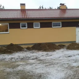 Żółta elewacja budynku z brązowym pasem, czerwony dach, trzy okna, dwoje drzwi i pryzmy ziemi na lekko ośnieżonym terenie.