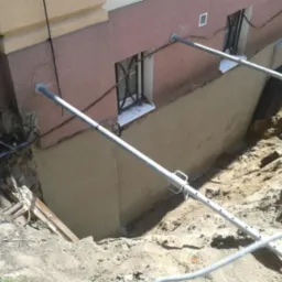 Odkopane fundamenty budynku z nową warstwą izolacyjną, wsparte metalowymi stemplami, widoczne okno piwniczne.