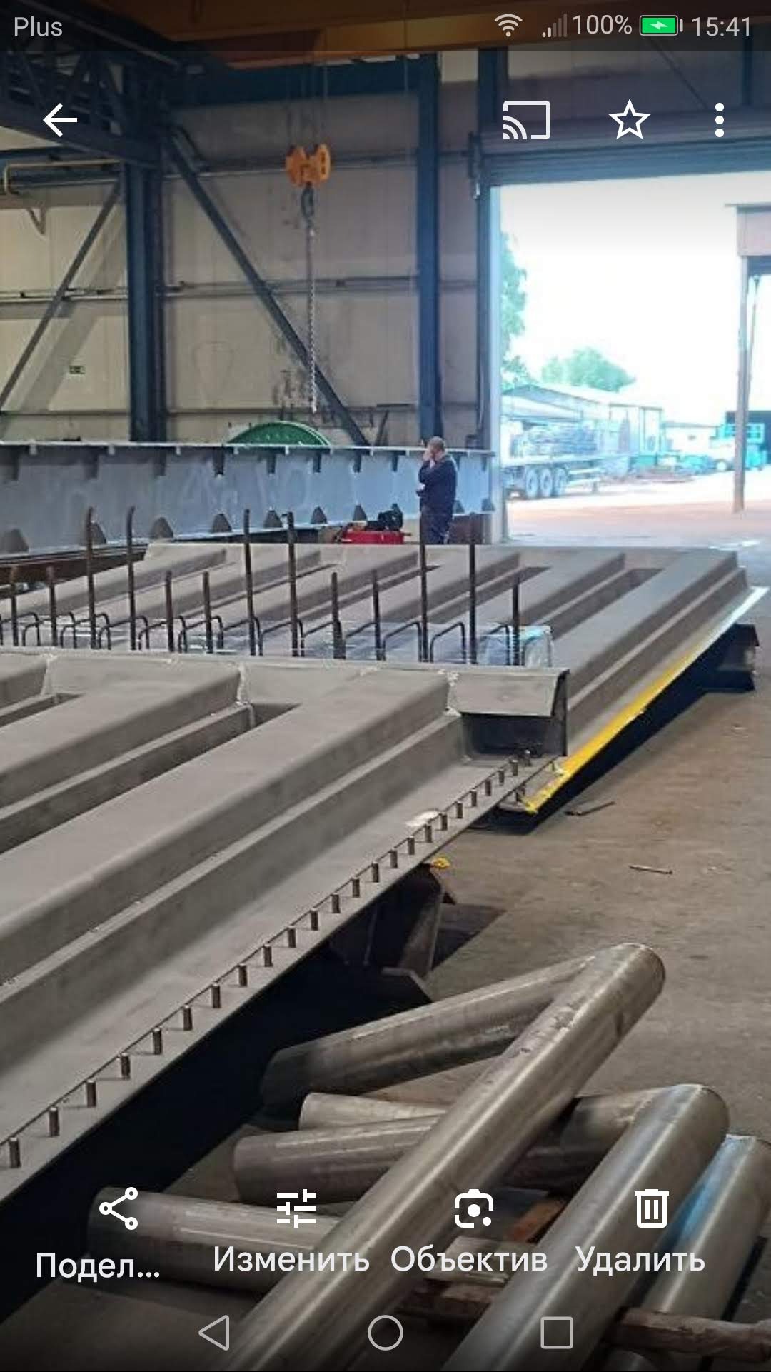 Wnętrze hali produkcyjnej z elementami konstrukcyjnymi, prawdopodobnie prefabrykatami betonowymi do budowy domu modułowego, z widocznym dźwigiem i pracownikiem w tle.