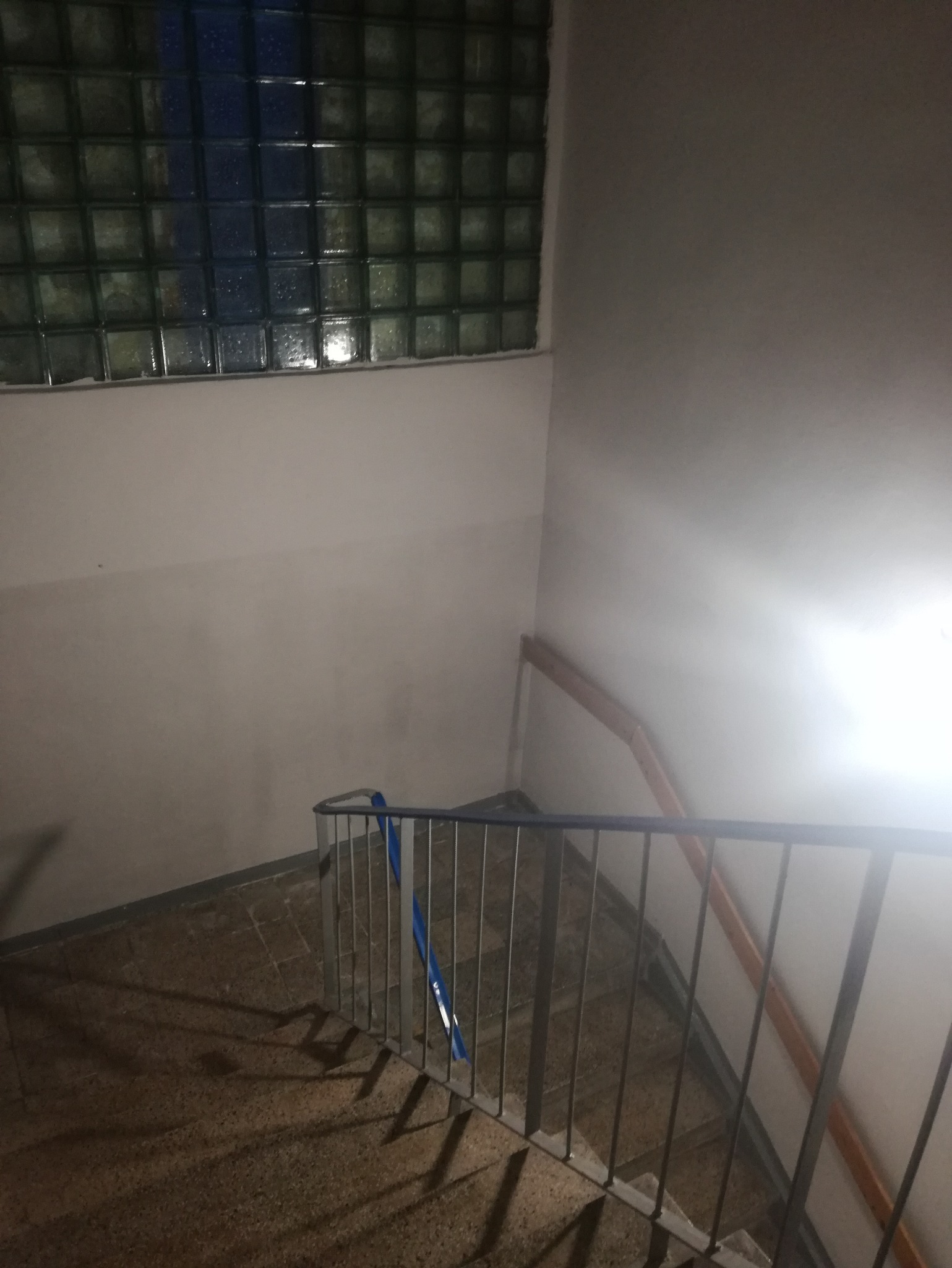 Klatka schodowa z oknem z luksferów. Metalowa barierka z niebieskim akcentem. Ściany w odcieniach bieli. Ujęcie z góry, widoczne schody i fragment poręczy.