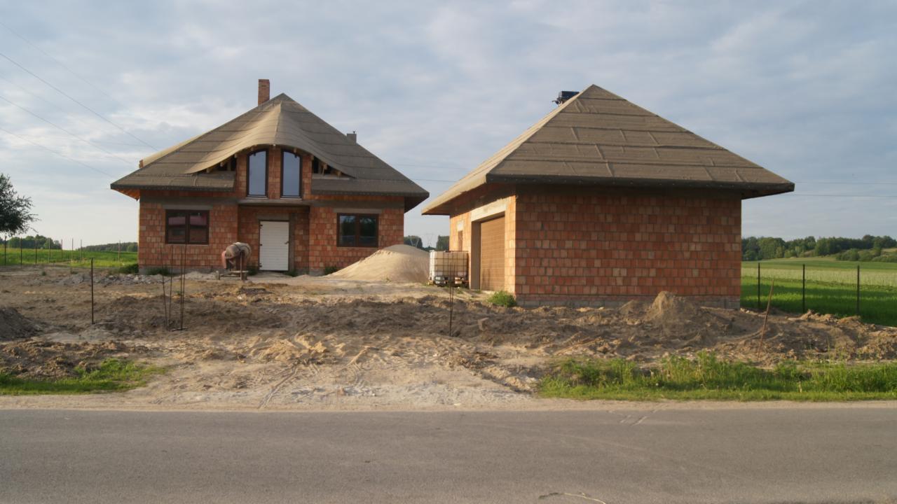 Budowa domu z czerwonej cegły i garażu z brązowym dachem. Na pierwszym planie droga i teren budowy z piaskiem. Widoczna osoba pracująca przy budynku.