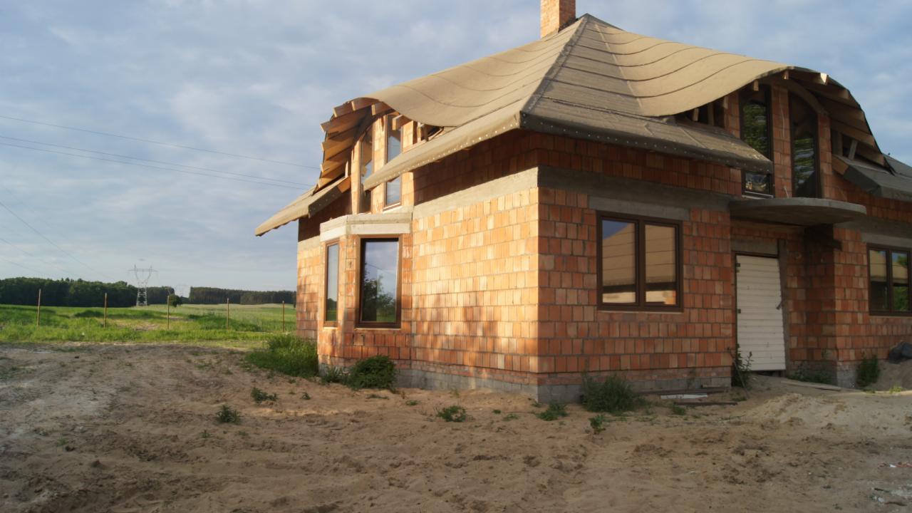 Niedokończony dom z cegły ceramicznej i częściowo ułożonym, falistym pokryciem dachowym. Widoczne okna w brązowych ramach. W tle łąka i las.