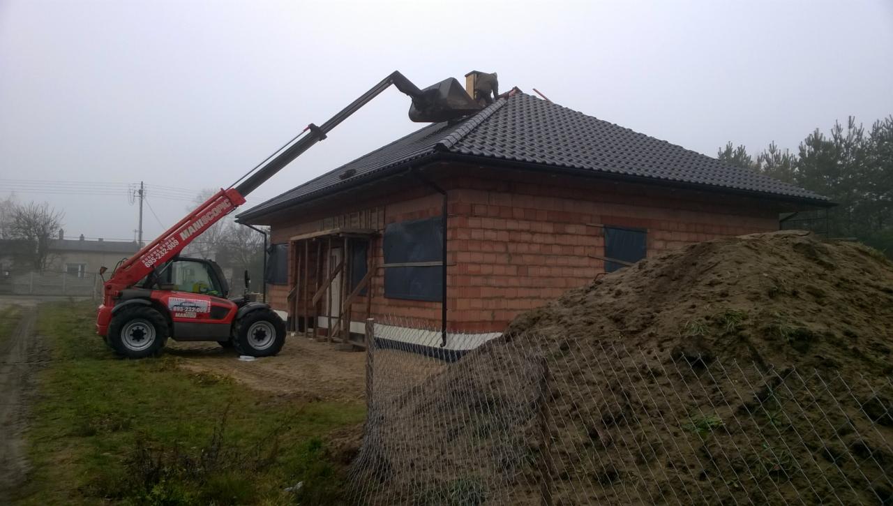 Układanie dachówki na nowym domu jednorodzinnym za pomocą podnośnika teleskopowego Maniscopic, widoczny mur z czerwonej cegły i sterta ziemi.
