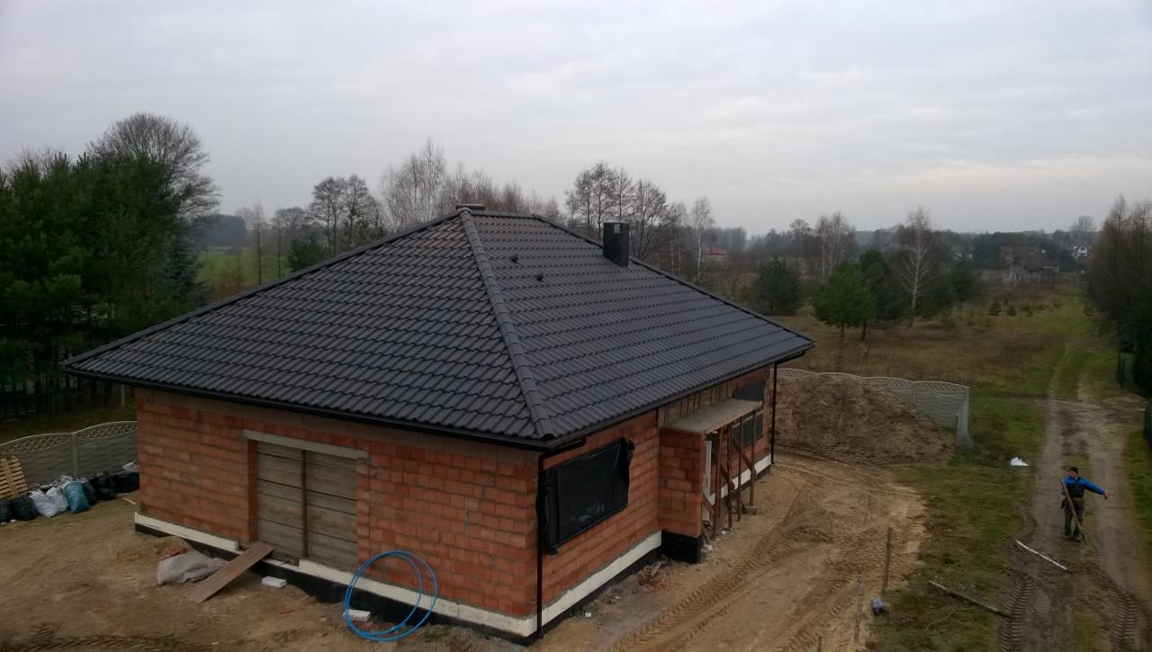 Dom w trakcie budowy z ciemną, nową dachówką. Widoczny komin, ceglana elewacja i otoczenie w stanie surowym. Całość w pochmurny dzień.