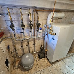 Fetta Fet-Dom - Nowoczesny piec gazowy z rozbudowaną instalacją hydrauliczną i zaworami, zamontowany w pomieszczeniu wyłożonym płytkami. Widoczne rury miedziane i elementy sterujące.