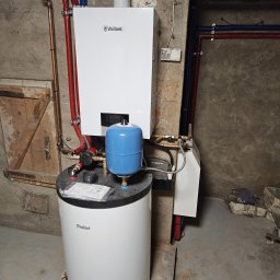 Fetta Fet-Dom - Biały piec gazowy Vaillant z niebieskim zbiornikiem wyrównawczym, zamontowany na tle surowej ściany piwnicy z widocznymi rurami instalacji grzewczej.