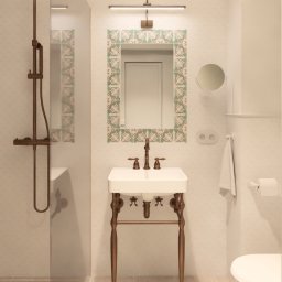 La Tua Isola Studio Julia Białaszewska - Biała umywalka na metalowym stelażu w łazience z kabiną prysznicową i lustrem w ozdobnej ramie z płytek. Styl retro, ciepłe oświetlenie.