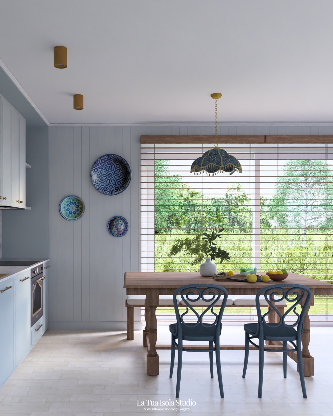 Jasna kuchnia w stylu rustykalnym z drewnianym stołem, niebieskimi krzesłami i dekoracyjnymi talerzami na ścianie. Widok na ogród przez okno z żaluzjami.