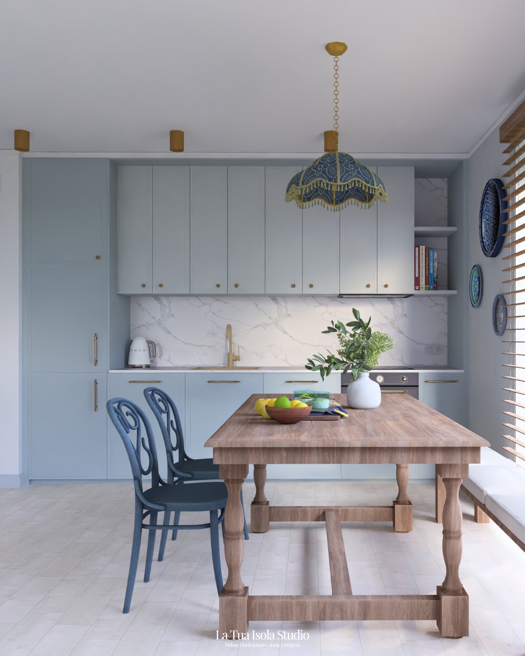 Jasna kuchnia z błękitnymi szafkami, marmurowym blatem i drewnianym stołem. Nad stołem wisi ozdobna lampa. Na stole owoce i wazon z zielonymi gałązkami.