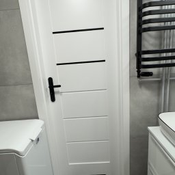 Fast Bilding Vasyl Voloshyn - Białe drzwi z czarnymi akcentami w nowoczesnej łazience z szarą płytką, grzejnikiem drabinkowym i białą szafką pod umywalką. Minimalistyczny design.