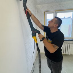 Fast Bilding Vasyl Voloshyn - Pracownik szlifuje ścianę szlifierką z odkurzaczem, przygotowując ją do malowania. Widoczne ślady po wcześniejszym wzorze na suficie i górnej części ściany.