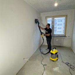 Fast Bilding Vasyl Voloshyn - Pracownik szlifuje ścianę w pokoju z wzorzystą tapetą, używając szlifierki z odkurzaczem. Widoczne narzędzia i przygotowane ściany do dalszych prac wykończeniowych.