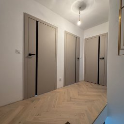 Beżowe drzwi z czarnymi akcentami w jasnym korytarzu z parkietem ułożonym w jodełkę. Widoczne trzy pary drzwi i fragment instalacji oświetleniowej na suficie.