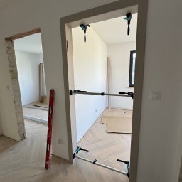 Fast Bilding Vasyl Voloshyn - Montaż ościeżnicy drzwiowej z użyciem rozpór i poziomicy w jasnym wnętrzu z parkietem w jodełkę. Widoczny fragment drugiego pomieszczenia w trakcie prac wykończeniowych.