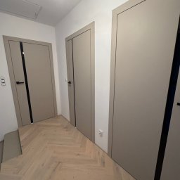 Fast Bilding Vasyl Voloshyn - Trzy beżowe drzwi wewnętrzne z czarnymi akcentami, widoczne na tle parkietu ułożonego w jodełkę, oświetlone górnym światłem. Minimalistyczny design.