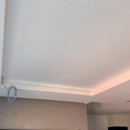 Fast Bilding Vasyl Voloshyn - Biały sufit z widocznymi śladami szpachlowania, fragment ściany z telewizorem i dwoma osobami w trakcie prac wykończeniowych. Widoczne kable przy suficie.