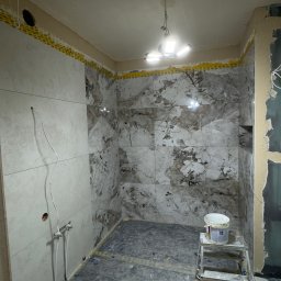 Fast Bilding Vasyl Voloshyn - Wnętrze łazienki w trakcie prac wykończeniowych: ułożone płytki imitujące marmur na ścianach, widoczne narzędzia glazurnicze i drabina z wiadrem farby.