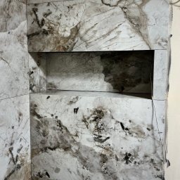 Fast Bilding Vasyl Voloshyn - Wykończona wnęka w ścianie wyłożonej płytkami imitującymi marmur. Widoczne fugi i narożnik. Biały kabel zwisa wzdłuż ściany.