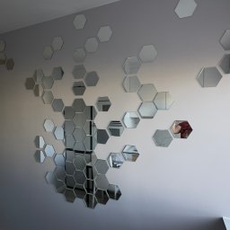 Fast Bilding Vasyl Voloshyn - Ściana w pokoju z dekoracyjnymi, lustrzanymi heksagonami tworzącymi wzór plastra miodu. Widoczny fragment gniazdka elektrycznego i odbicie osoby w jednym z luster.