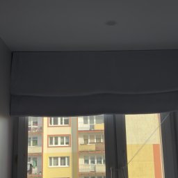 Fast Bilding Vasyl Voloshyn - Zamknięta roleta rzymska w jasnym odcieniu, zamontowana w oknie z widokiem na blok mieszkalny. Dekoracyjne, geometryczne elementy na ścianie po lewej stronie.