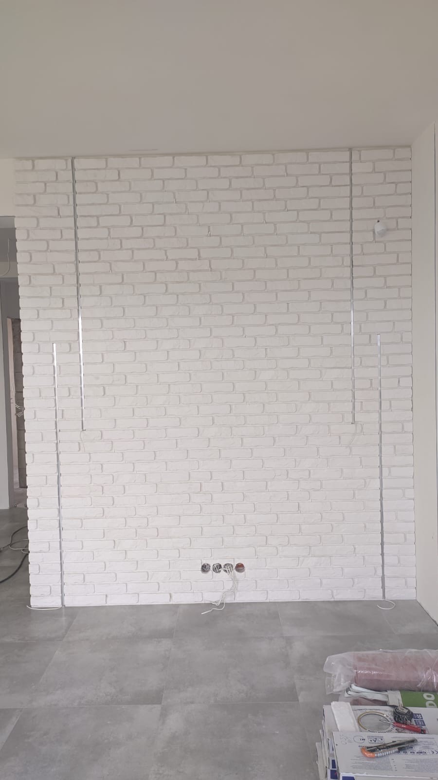 Ściana z białej cegły dekoracyjnej z metalowymi listwami w Stargardzie. Widoczne gniazdka elektryczne i szara podłoga. Minimalistyczny design wnętrza.