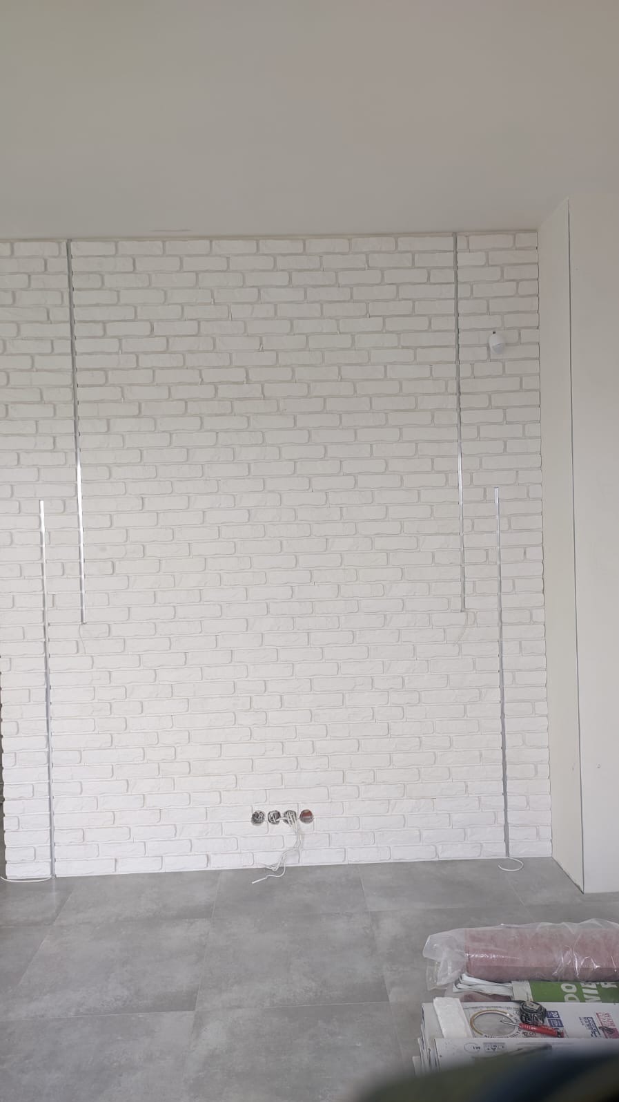 Biała ściana z cegły dekoracyjnej z listwami montażowymi, widoczne gniazdka elektryczne i szara podłoga. Minimalistyczne wykończenie wnętrza w jasnych barwach.