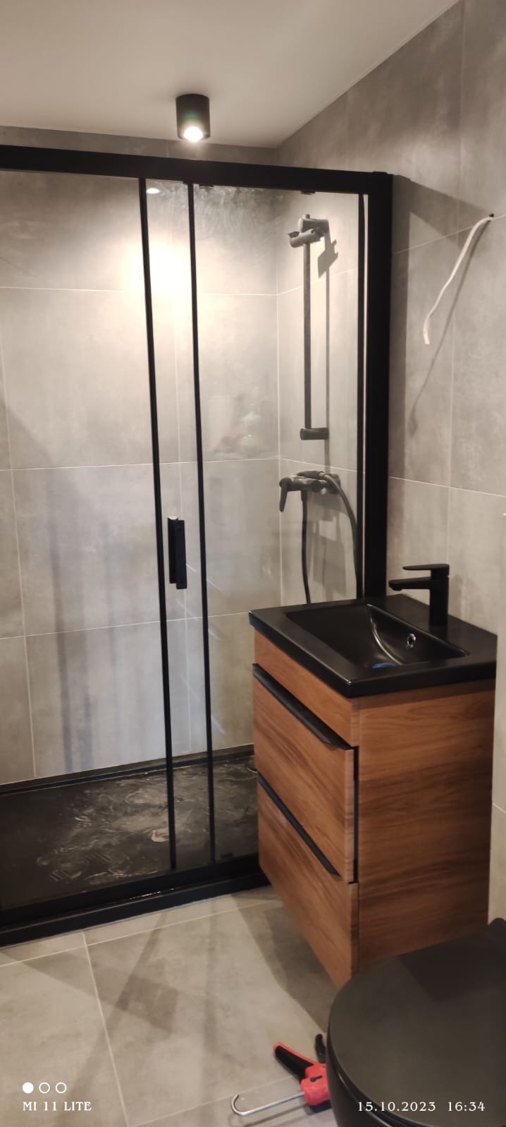 Nowoczesna łazienka z kabiną prysznicową typu walk-in z czarnymi profilami, drewnianą szafką pod umywalkę i szarą płytką. Widoczny silikon na podłodze.