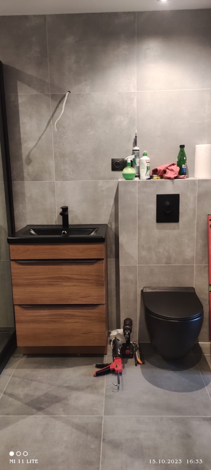 Nowoczesna łazienka w szarościach z drewnianą szafką pod umywalką i wiszącą toaletą. Widoczne narzędzia sugerują trwający montaż.