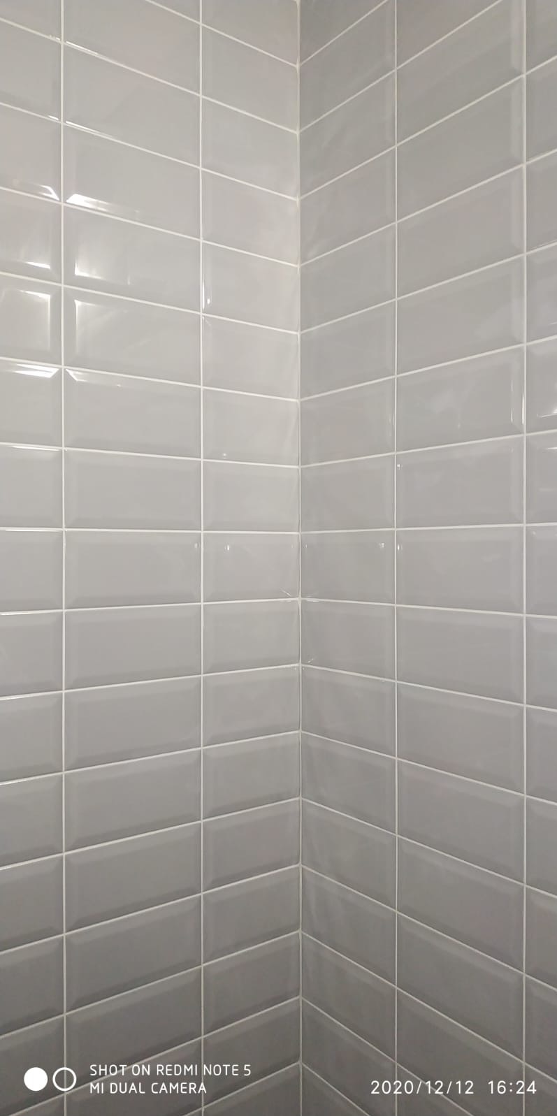 Ściana wyłożona szarymi, błyszczącymi, prostokątnymi płytkami ceramicznymi z białą fugą, tworzącymi efektowny, nowoczesny wzór w rogu pomieszczenia. Widoczne refleksy światła.