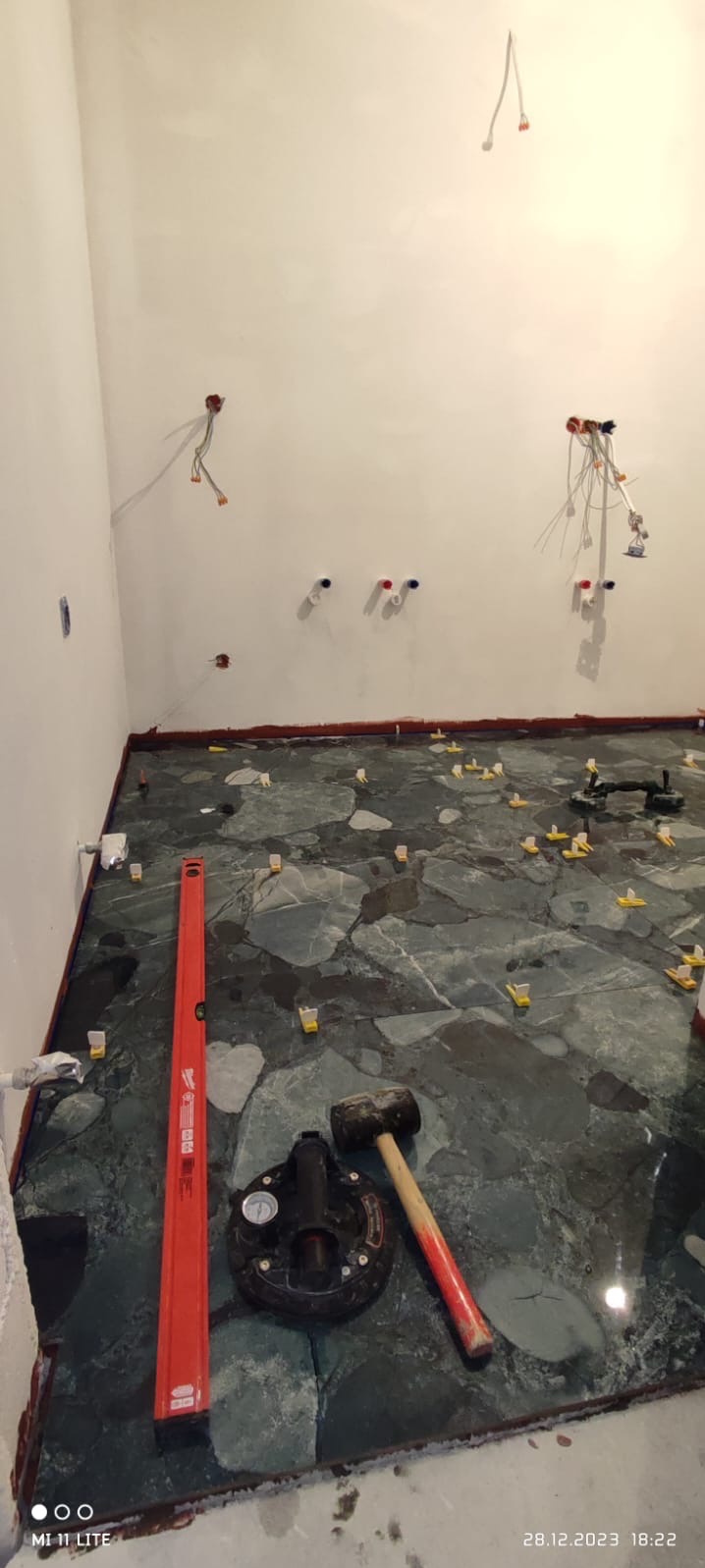 Układanie płytek podłogowych w łazience z użyciem poziomicy, przyssawki i młotka. Widoczne kable elektryczne i rury wodne w ścianie. Płytki w odcieniach zieleni i szarości.