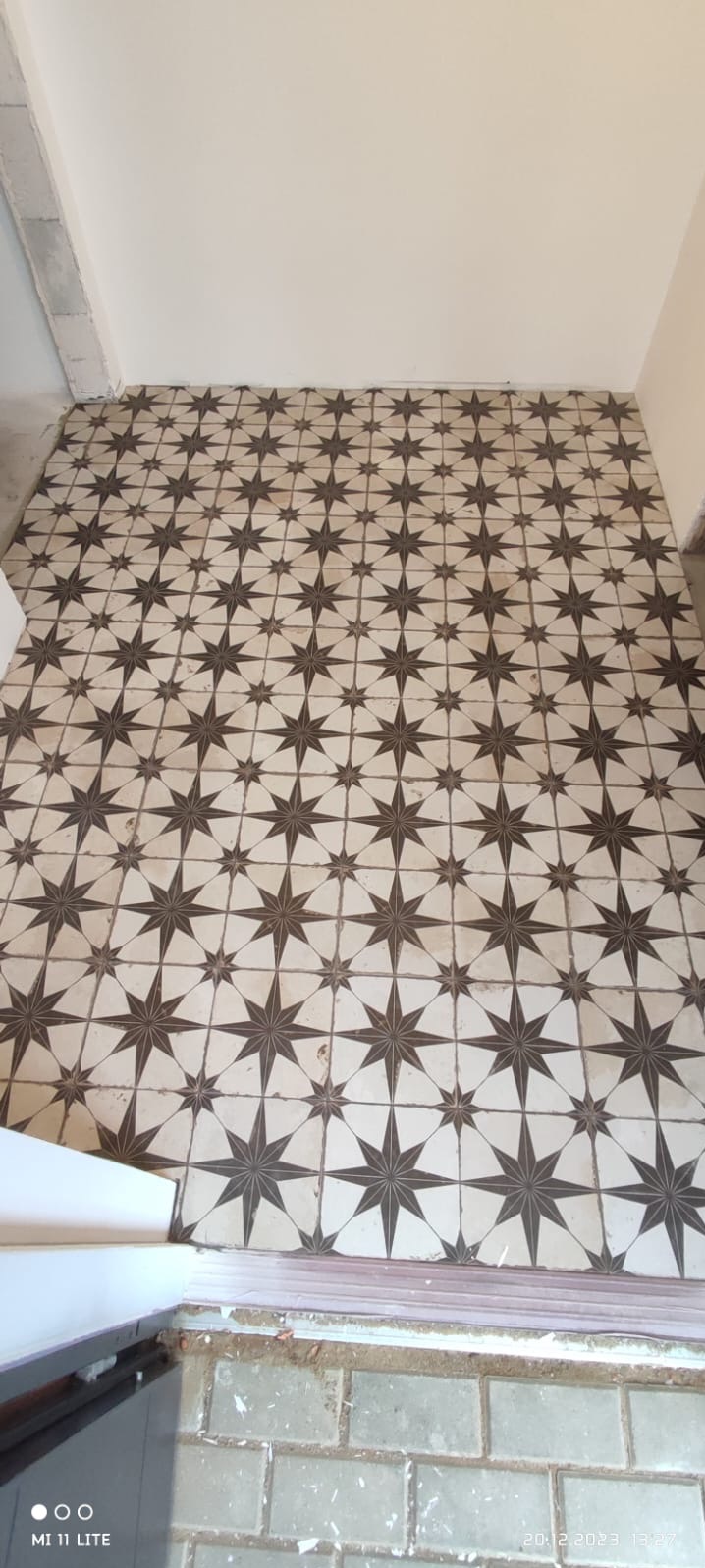 Ułożona podłoga z płytek w kremowo-brązowe gwiazdy, widok z góry. Płytki ułożone w regularny wzór, widoczne fugi. Fragment ściany w tle, surowy stan wykończenia.