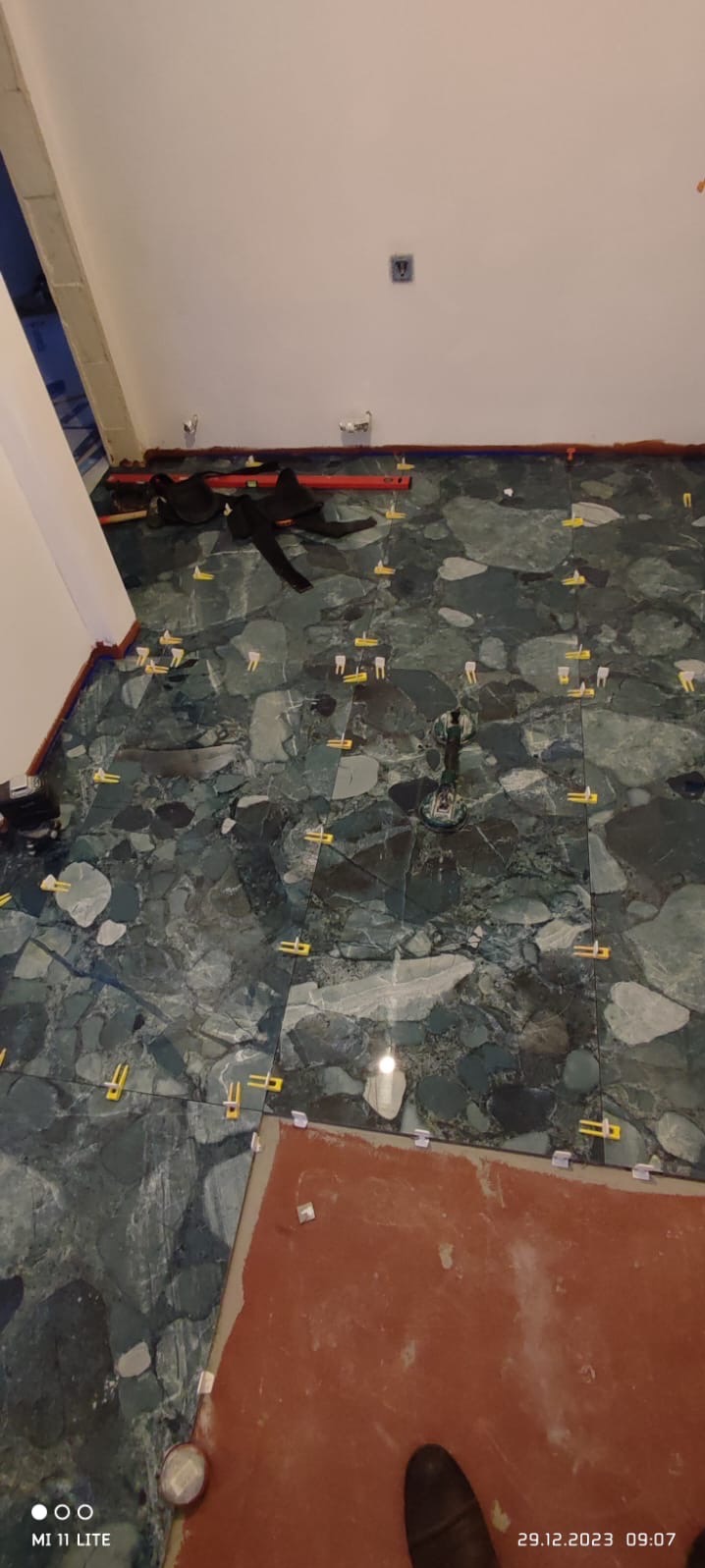 Układanie płytek podłogowych z użyciem poziomicy i klipsów dystansowych. W tle widoczna ściana i fragment niezakończonej podłogi.