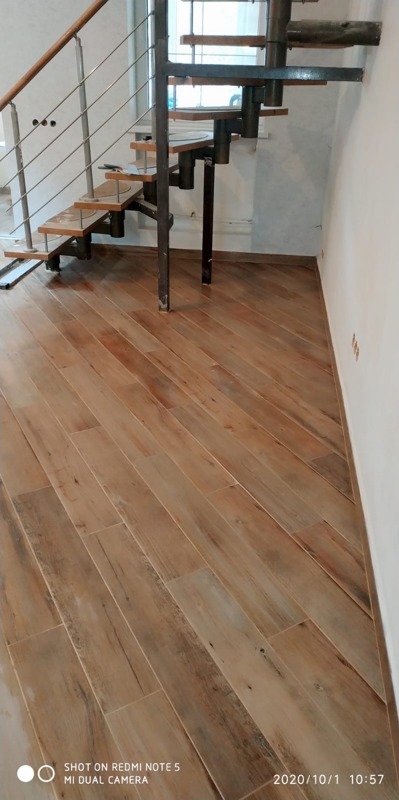 Ukończona podłoga z płytek imitujących drewno, ułożonych pod kątem, z widocznymi schodami spiralnymi w tle. Jasne ściany i naturalne światło podkreślają ciepły odcień podłogi.