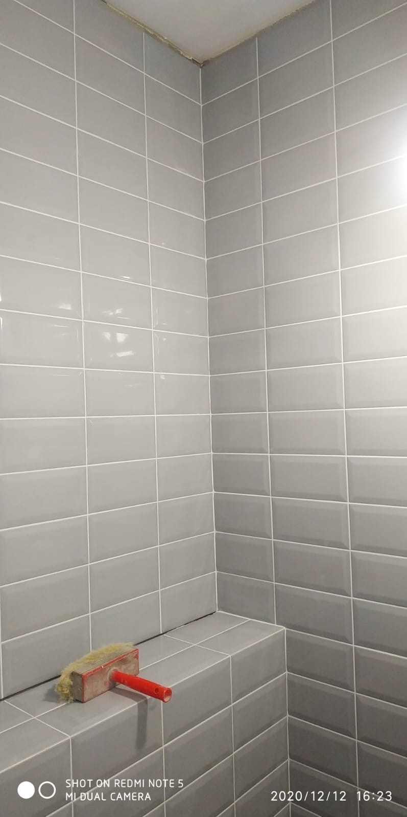 Wykończona łazienka z szarymi, błyszczącymi płytkami ułożonymi w regularny wzór. Na półce leży pędzel do fugowania z czerwoną rączką. Widoczny narożnik i fragment sufitu.
