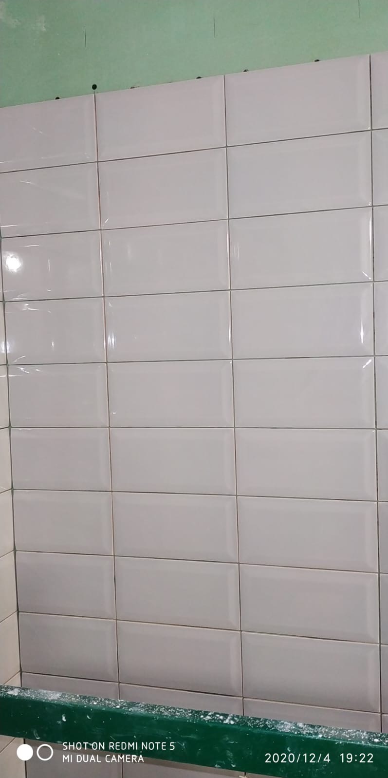 Ściana wyłożona białymi, błyszczącymi płytkami w kształcie prostokątów z ciemną fugą. Widoczny fragment zielonej ściany i krawędź wanny.