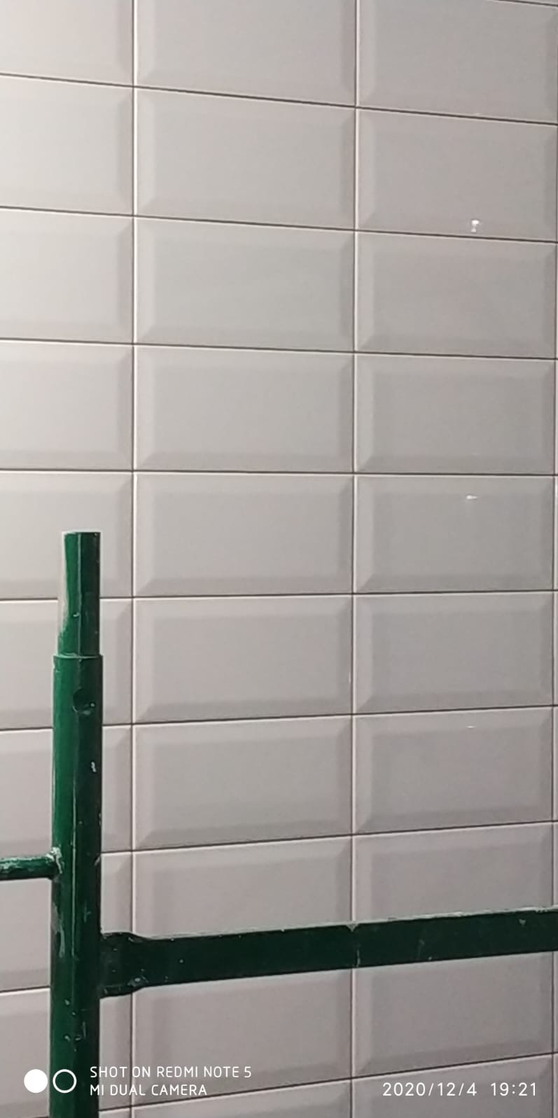 Ściana wyłożona białymi, prostokątnymi płytkami typu metro z widocznym fragmentem zielonego rusztowania. Ujęcie z bliska, ukazujące detale fug i połysk płytek.