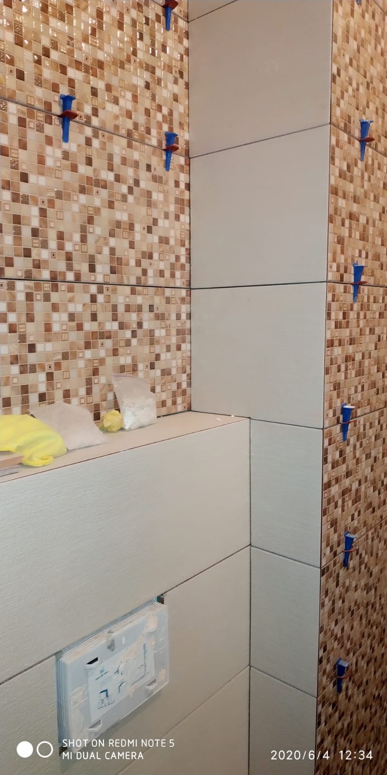 Układanie płytek w łazience: mozaika w odcieniach brązu i beżu kontrastuje z dużymi, jasnymi płytkami. Widoczne niebieskie kliny do poziomowania i akcesoria glazurnicze.