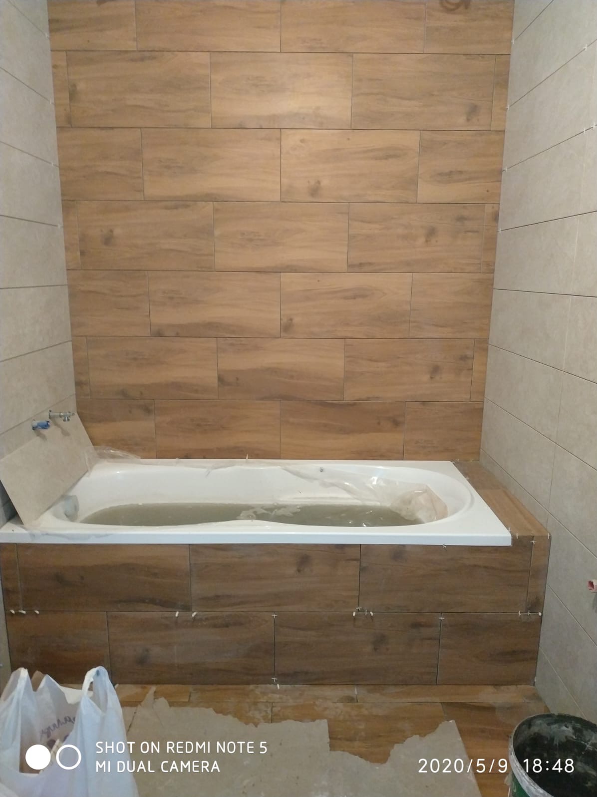 Łazienka w trakcie wykończenia: brązowe płytki imitujące drewno na ścianach i obudowie wanny, biała wanna zabezpieczona folią, widoczne krzyżyki glazurnicze.