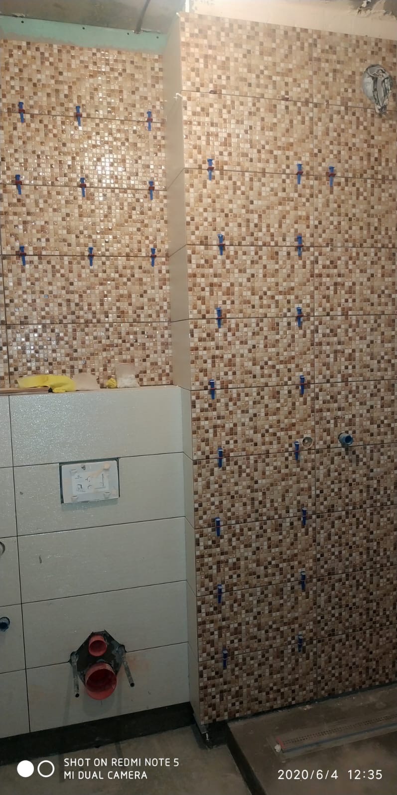 Mozaika w odcieniach brązu na ścianie łazienki, z niebieskimi klipsami poziomującymi. Częściowo wykończona ściana z jasnych płytek. Widoczne elementy instalacji wodnej.