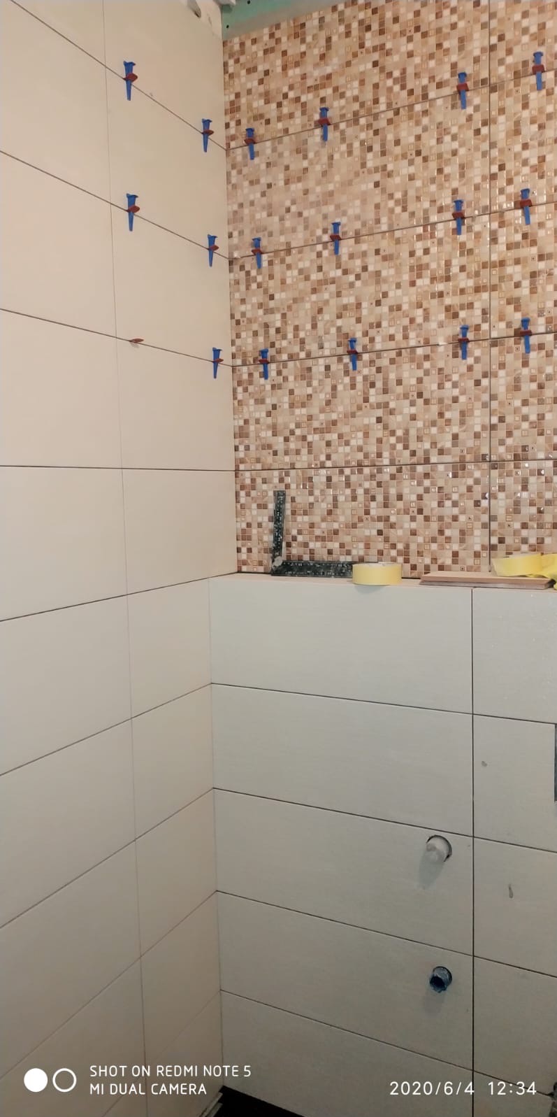 Układanie mozaiki i białych płytek w łazience. Widoczne niebieskie krzyżyki dystansowe i poziomica. Częściowo wykończona ściana, detale montażu.