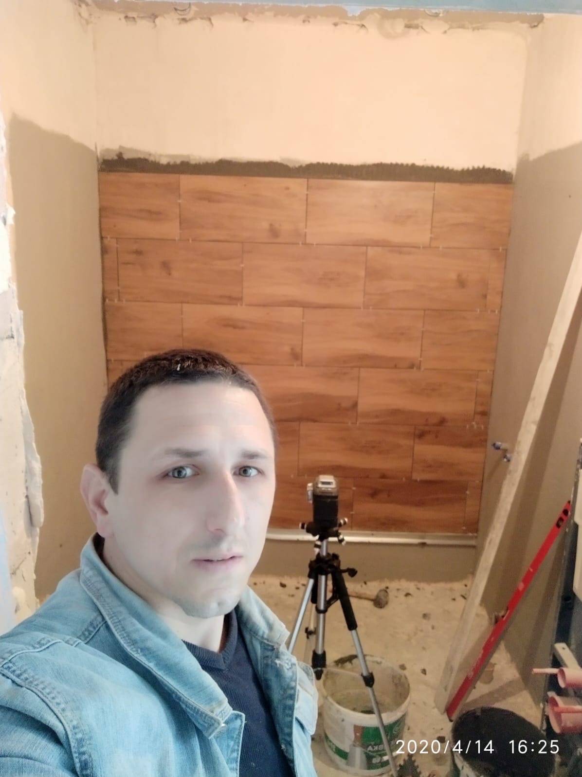 Ściana w trakcie układania płytek imitujących drewno, widoczny laser krzyżowy na statywie, poziomica i osoba w kadrze. Prace glazurnicze w łazience.