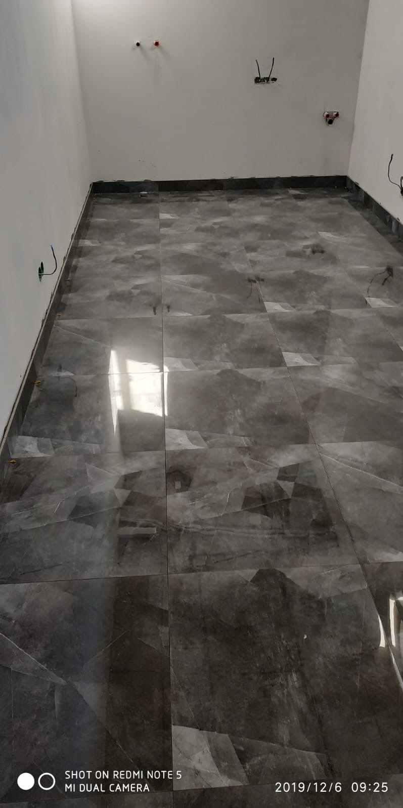 Ułożona, błyszcząca, szara podłoga z płytek imitujących marmur w pustym pomieszczeniu z wystającymi kablami elektrycznymi. Widoczne fugi i listwy przypodłogowe.